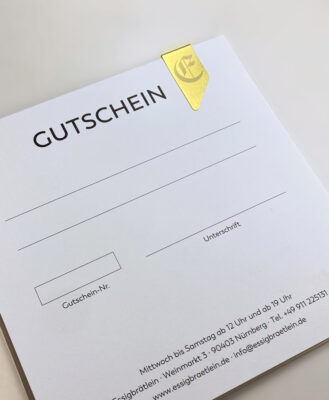 Essigbrätlein Gutschein: Eine goldene Papierklammer mit dem Initial-"E" des Logos hält den Gutschein in seiner Hülle.