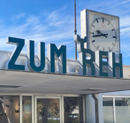 Die Ladenbeschriftung "Zum Reh" in großen Buchstaben in dunkelblau, im Hintergrund eine alte Bahnhofsuhr und blauer Himmel.